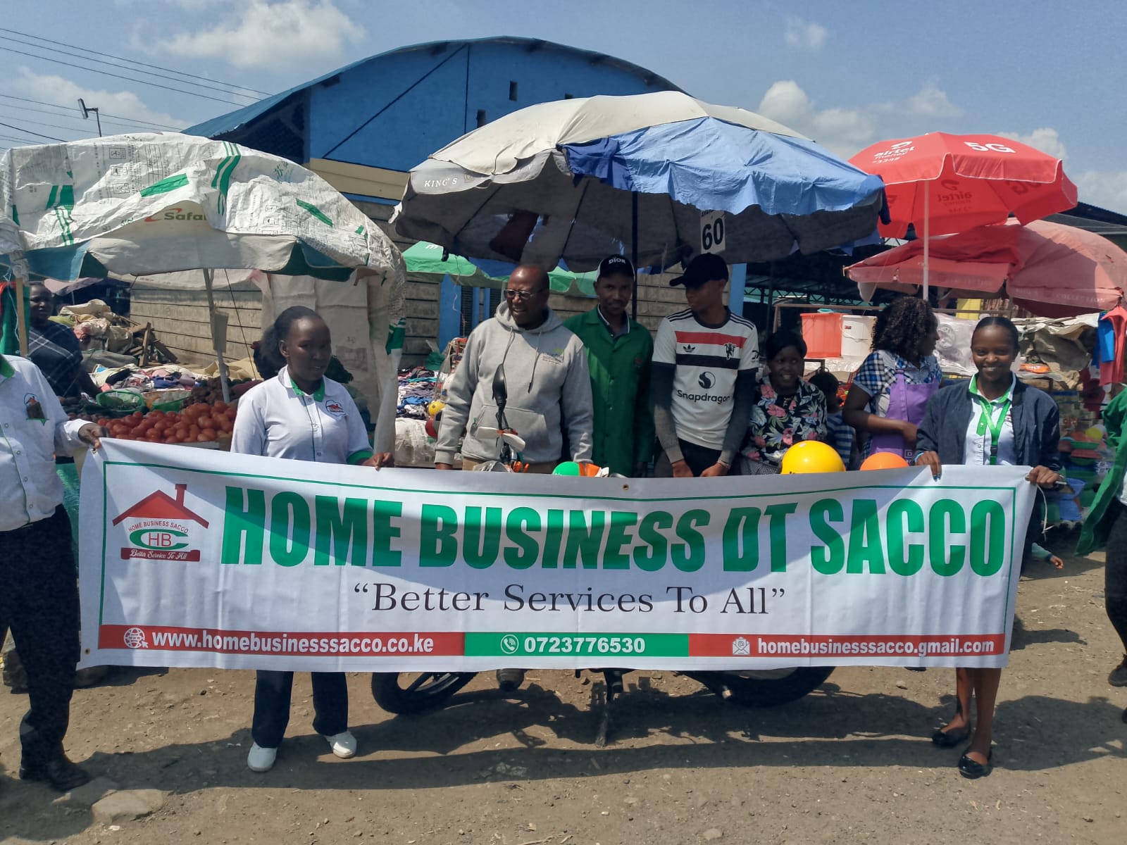 Group of people gathered around a red motorcycle decorated with a large orange bow, standing outdoors on a sunny day in front of shops with signs including Home Business DT Sacco Office and Joel Peters Milk Bar. The mood is celebratory and supportive.